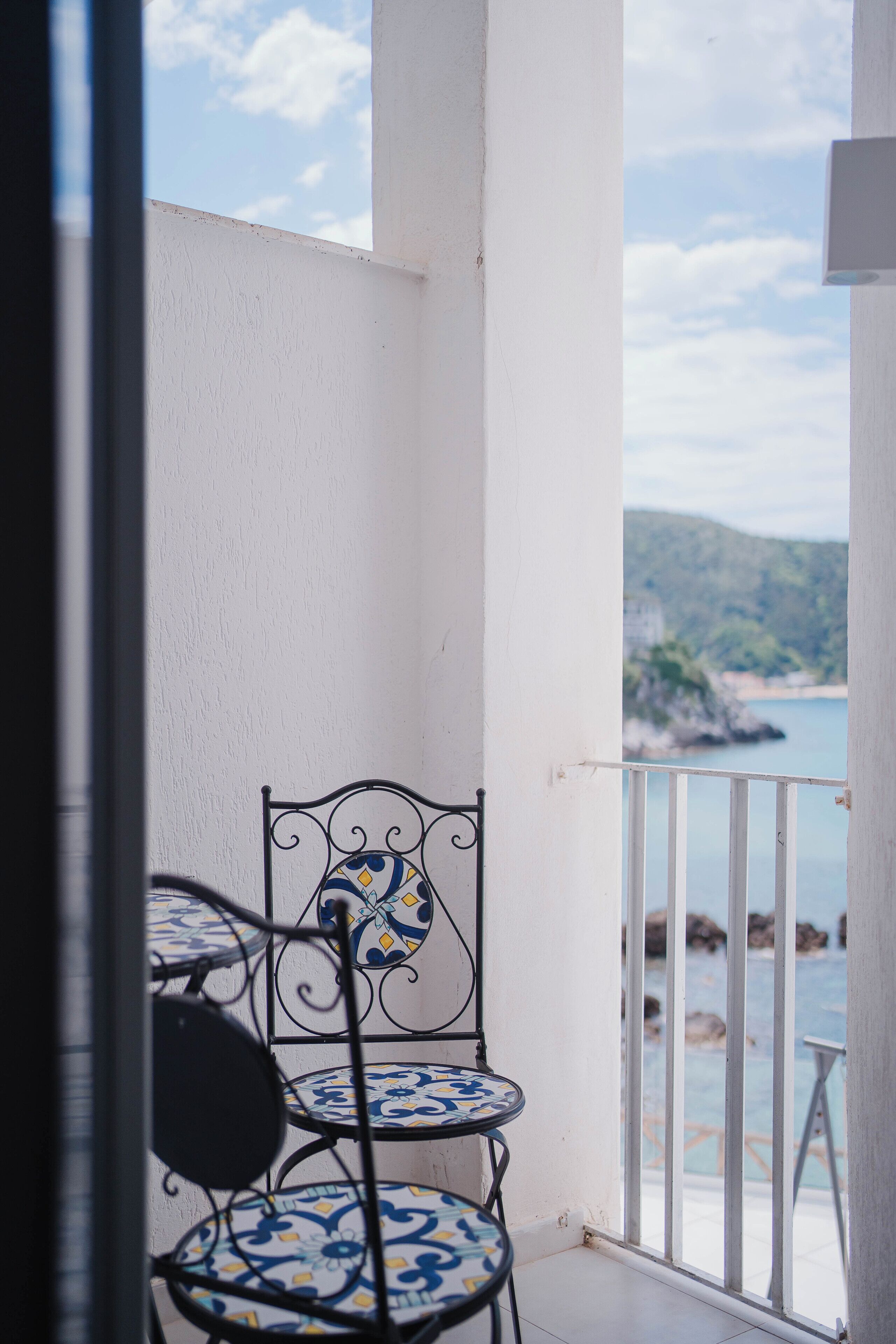 Un balcon avec une balustrade blanche, une chaise en métal avec un siège à motifs bleus et blancs, et une vue sur un paysage côtier.