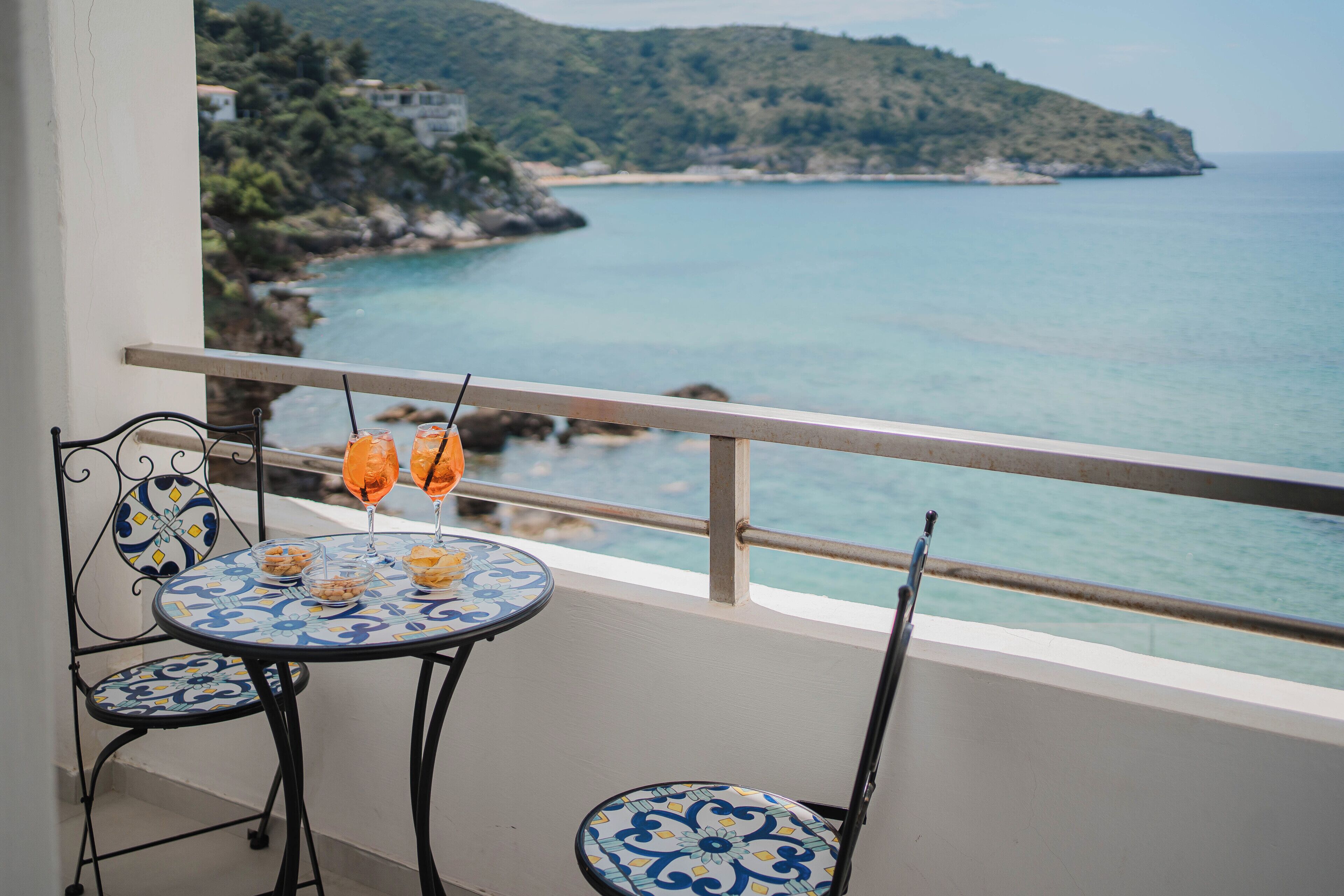 A balcony with two chairs and a table, overlooking a coastal view with buildings and greenery.