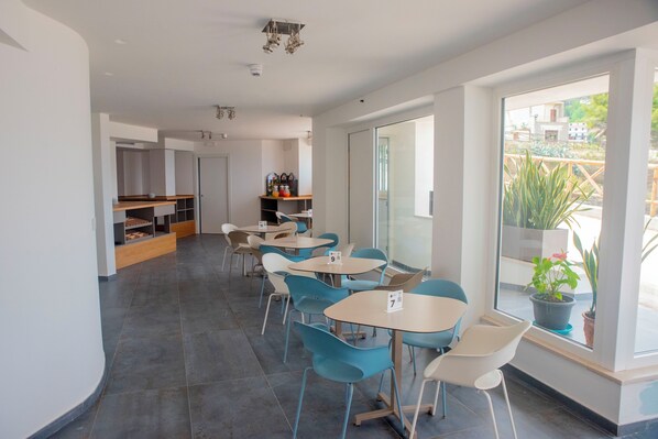 A modern interior with multiple tables and chairs, a visible door leading to another room, and a large window with a view of the outside. - Hotel Ficocella (Centola)