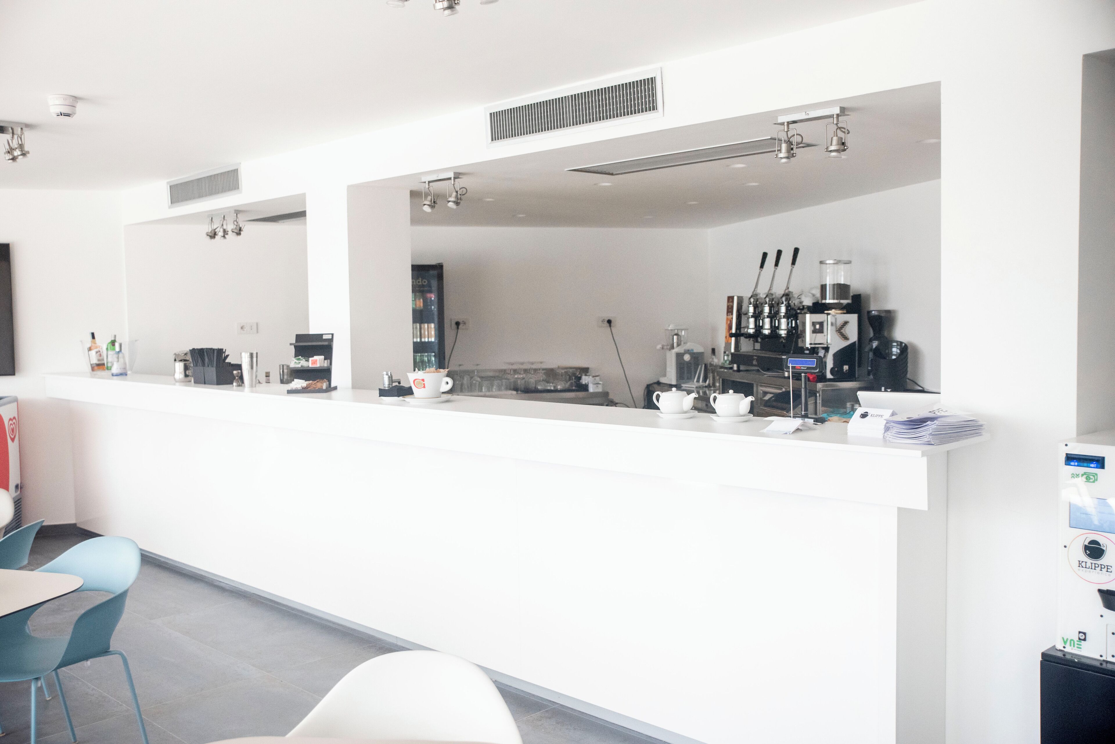 Una cafetería moderna y bien iluminada, con un interior limpio y blanco, una barra con equipo para preparar café y sillas azules.