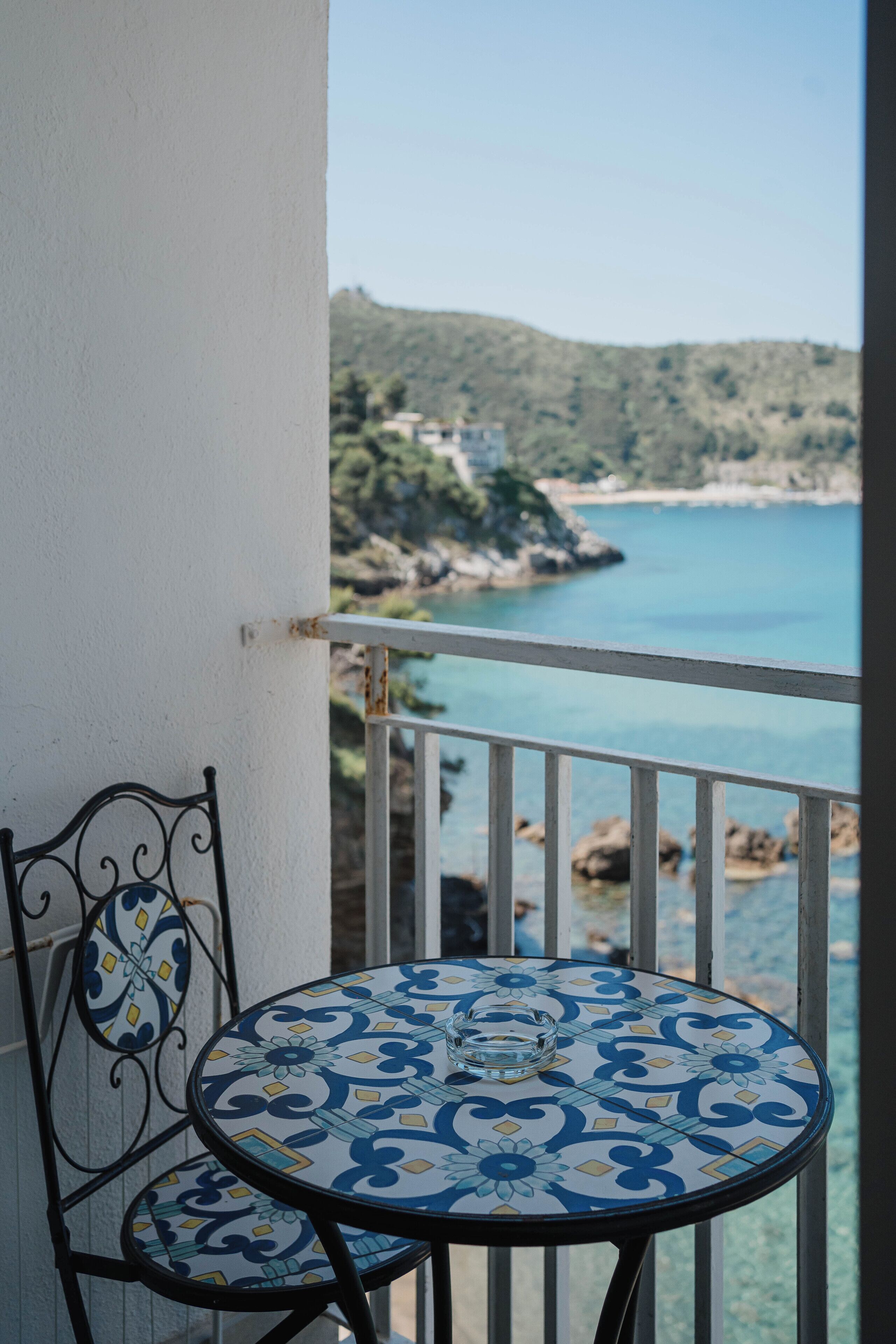 A balcony with a table and chair, overlooking a coastal landscape with hills and a body of water.