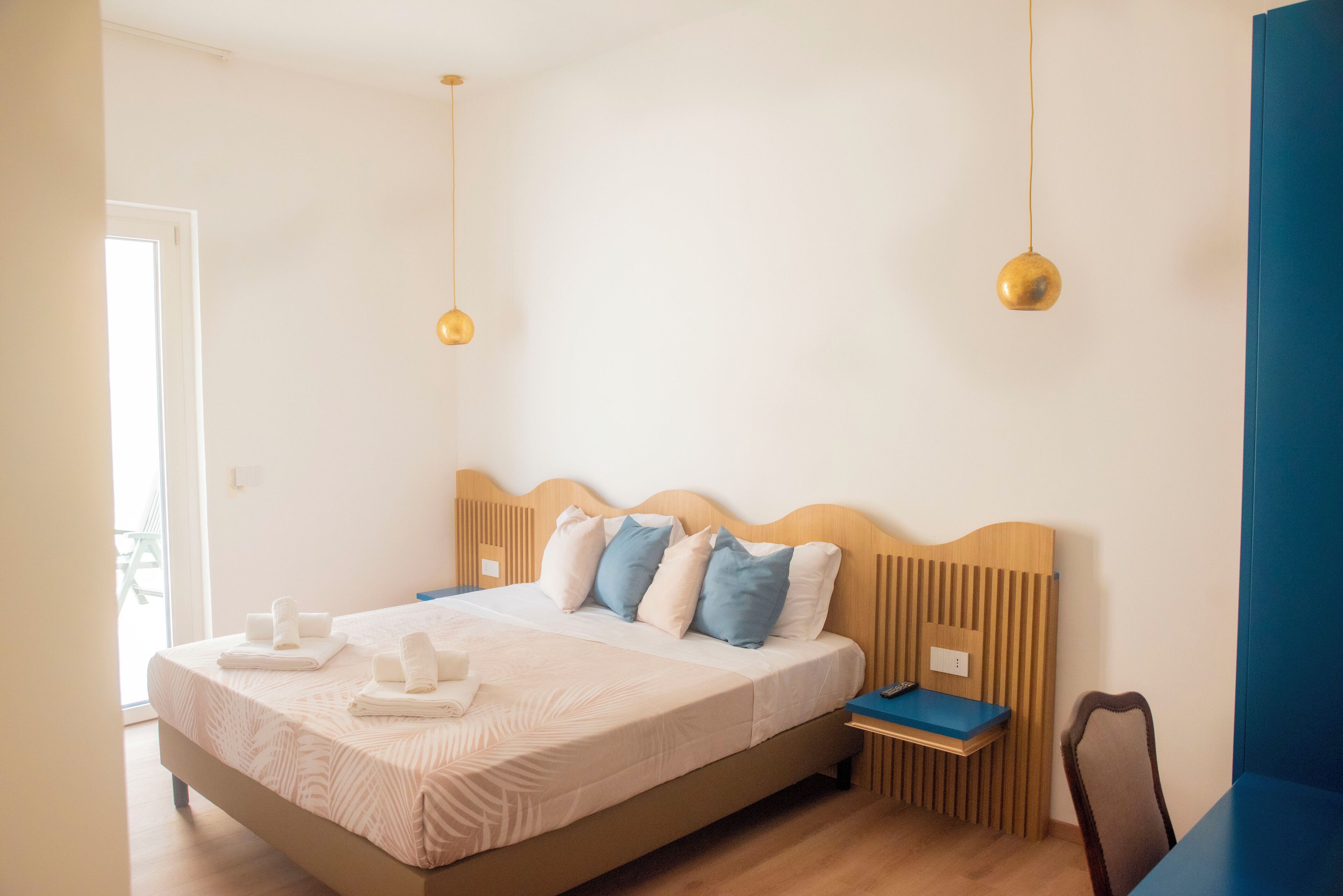 A neatly made bed with pillows, a bedside table, and a chair in a well-lit room with a wooden headboard and a hanging light fixture.