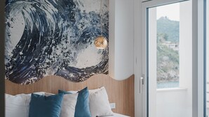 A bedroom with a large wave painting on the wall, a bed with blue and white bedding, and a view of the outdoors through a glass door. - Hotel Ficocella (Centola)