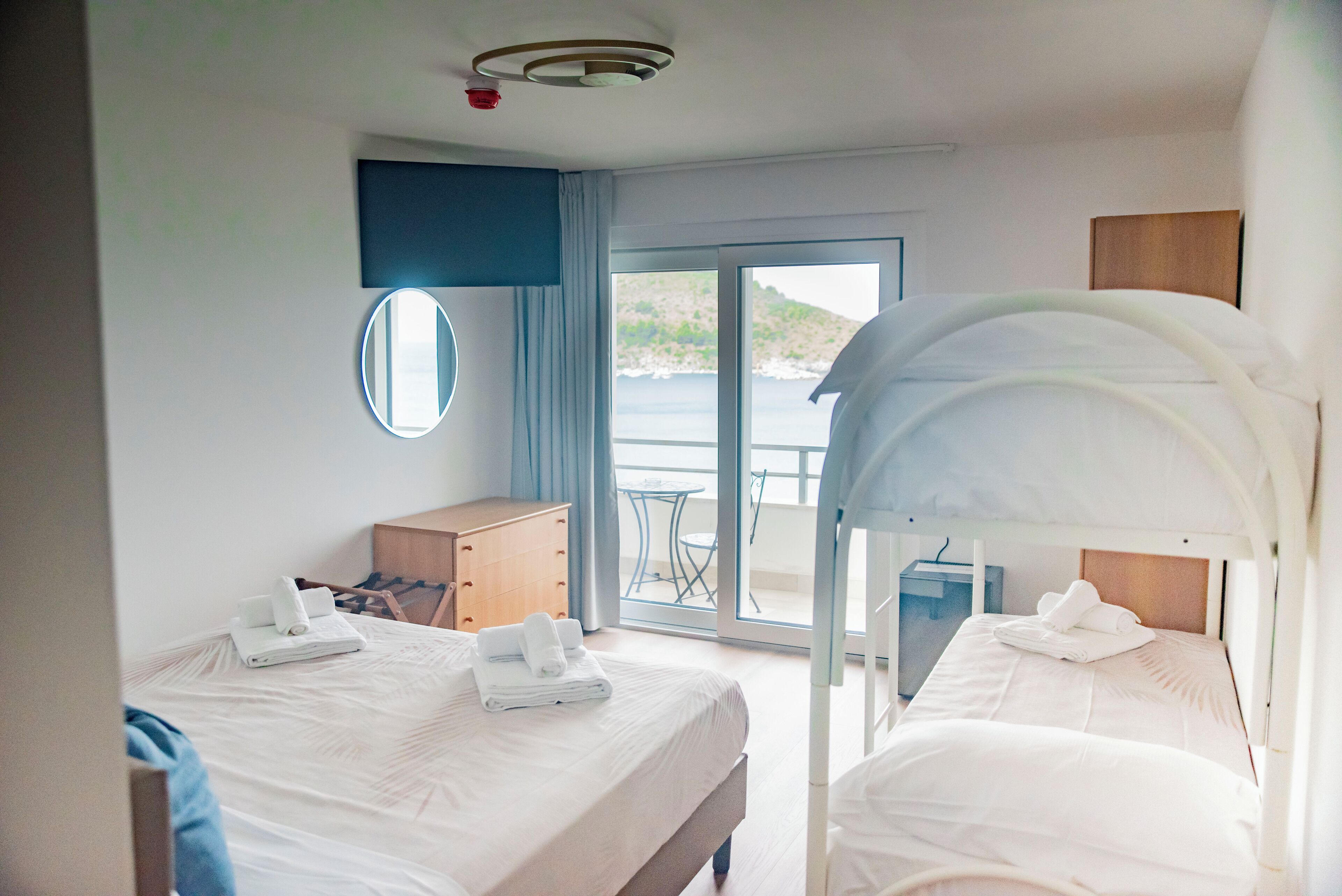 A hotel room with two beds, a balcony, a view of the sea, a wooden dresser, and a round mirror.