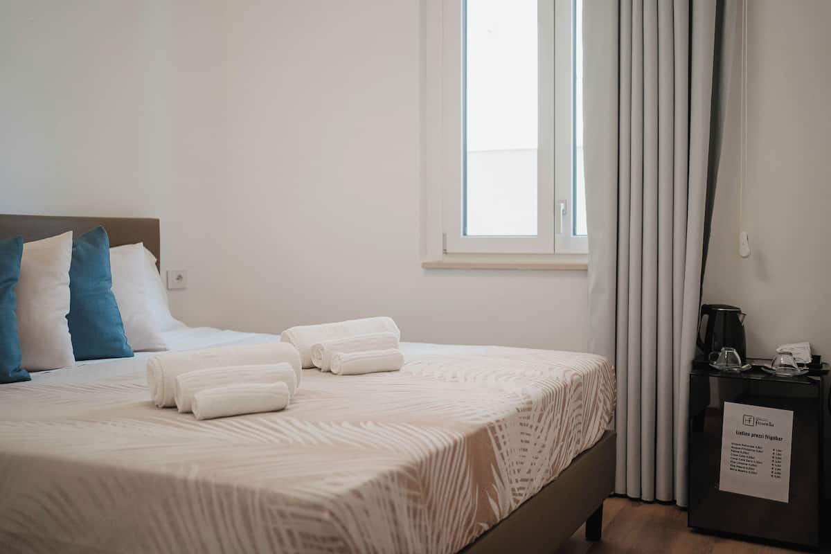 Ein ordentlich bezogenes Bett mit weißen und blauen Kissen, ein Fenster mit Vorhängen sowie ein Nachttisch mit Wasserkocher und einem Schild.