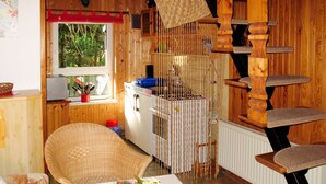 Una habitación acogedora con una silla de mimbre, una mesa con un centro de mesa floral, un rascador de madera para gatos y una ventana con vistas a la vegetación.