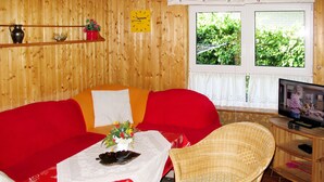 A cozy room with a wooden interior, a red sofa, a wicker chair, a small table with a white tablecloth, a television, and a window with a view of greenery. - Katarina I by Interhome (Plau am See)