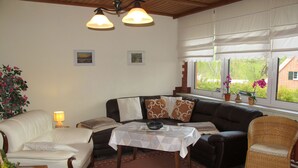 En hyggelig stue med sofa, lænestole, spisebord og en fletstol. Der er to pendellamper, et vindue med gardiner og en potteplante.
