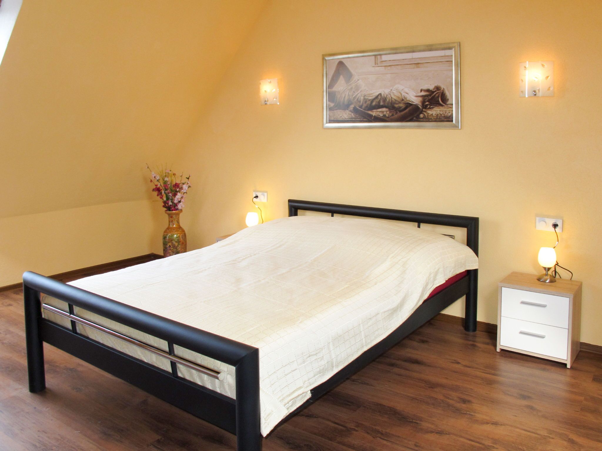 Un dormitorio con cama, mesita de noche, un jarrón con flores y un cuadro en la pared.