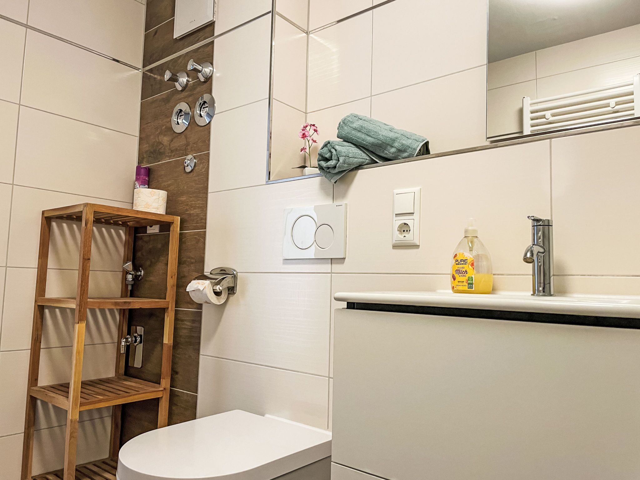 A modern bathroom with a toilet, a wooden shelf, a towel rack, and a soap dispenser.