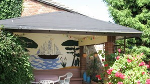 Une terrasse couverte ornée d’une fresque représentant un navire sur le mur, avec des chaises blanches et des plantes en pot.