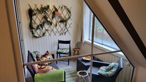 Een knus ingerichte kamer met een bed, een stoel en een bank. Er is een raam met gordijnen, een decoratief wandobject en een klein tafeltje met een kaars.