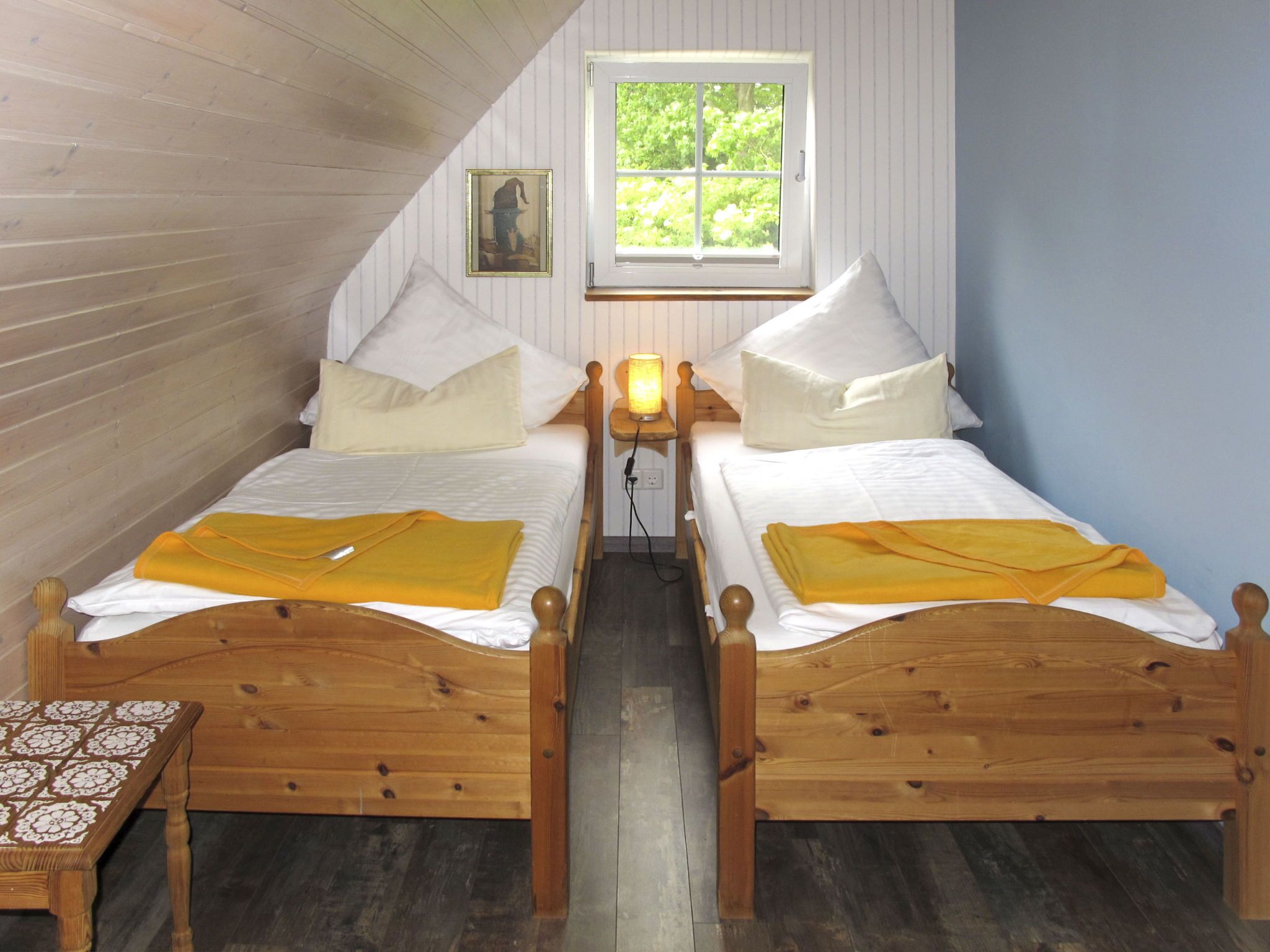 A room with two wooden beds, a window with a view of greenery, a small table, and a wall-mounted picture.