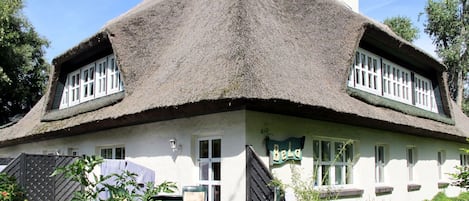 A traditional thatched-roof house with a white exterior, two windows with white frames, and a sign that reads "Bela".