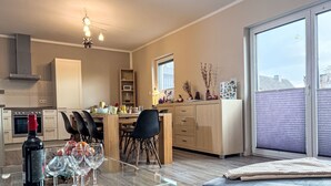 A modern kitchen with a dining area, featuring a glass table with wine glasses, a sofa, and a large window with blinds. - Johannika by Interhome (Godern)