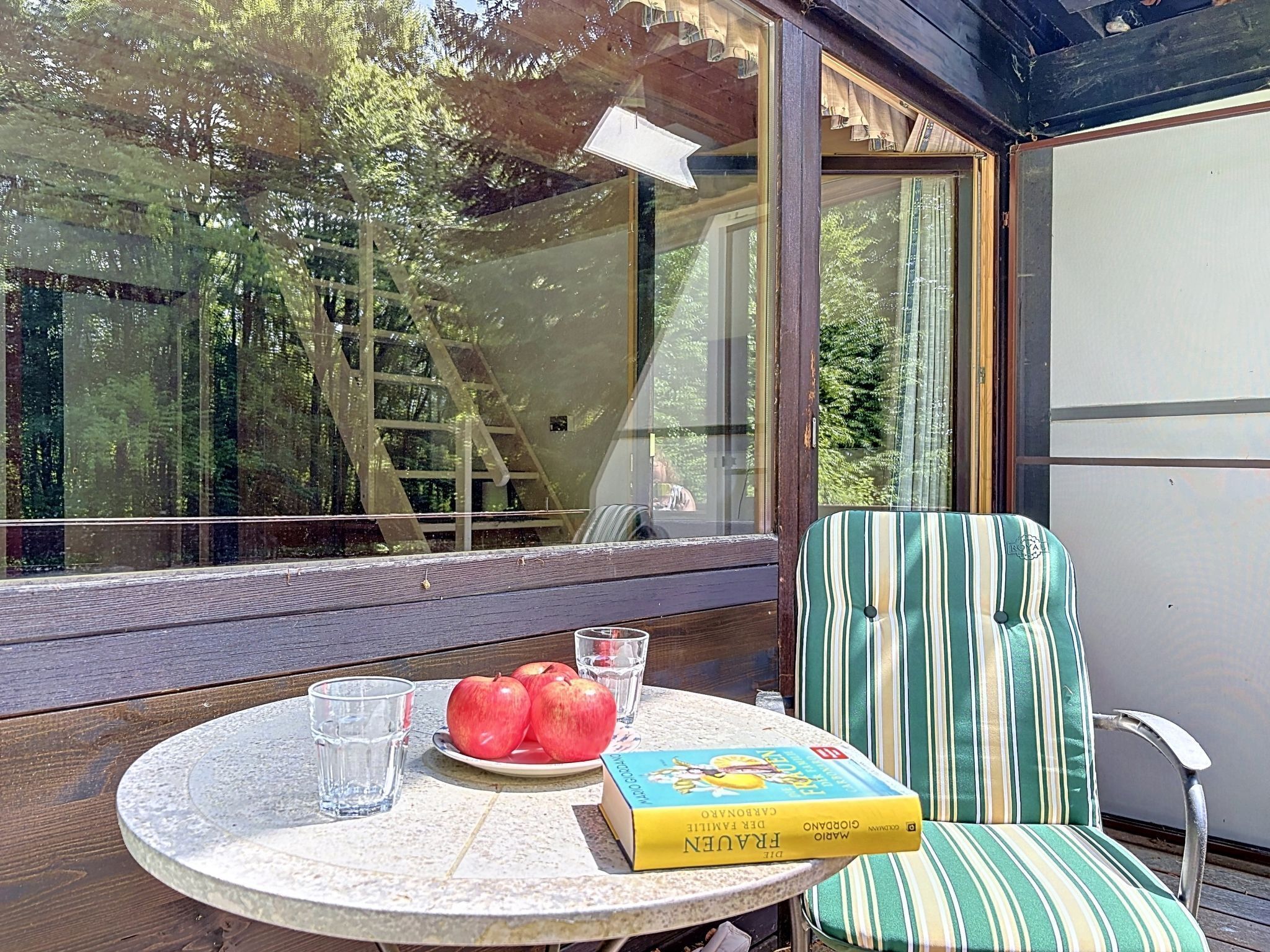 Una mesa redonda con un libro, manzanas y gafas, una silla a rayas y vistas de árboles a través de una ventana.