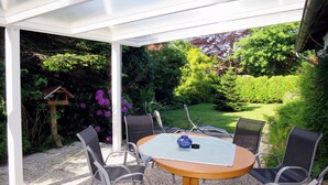 Une terrasse couverte avec une table ronde et six chaises, entourée d’une végétation luxuriante et offrant une vue sur le jardin.