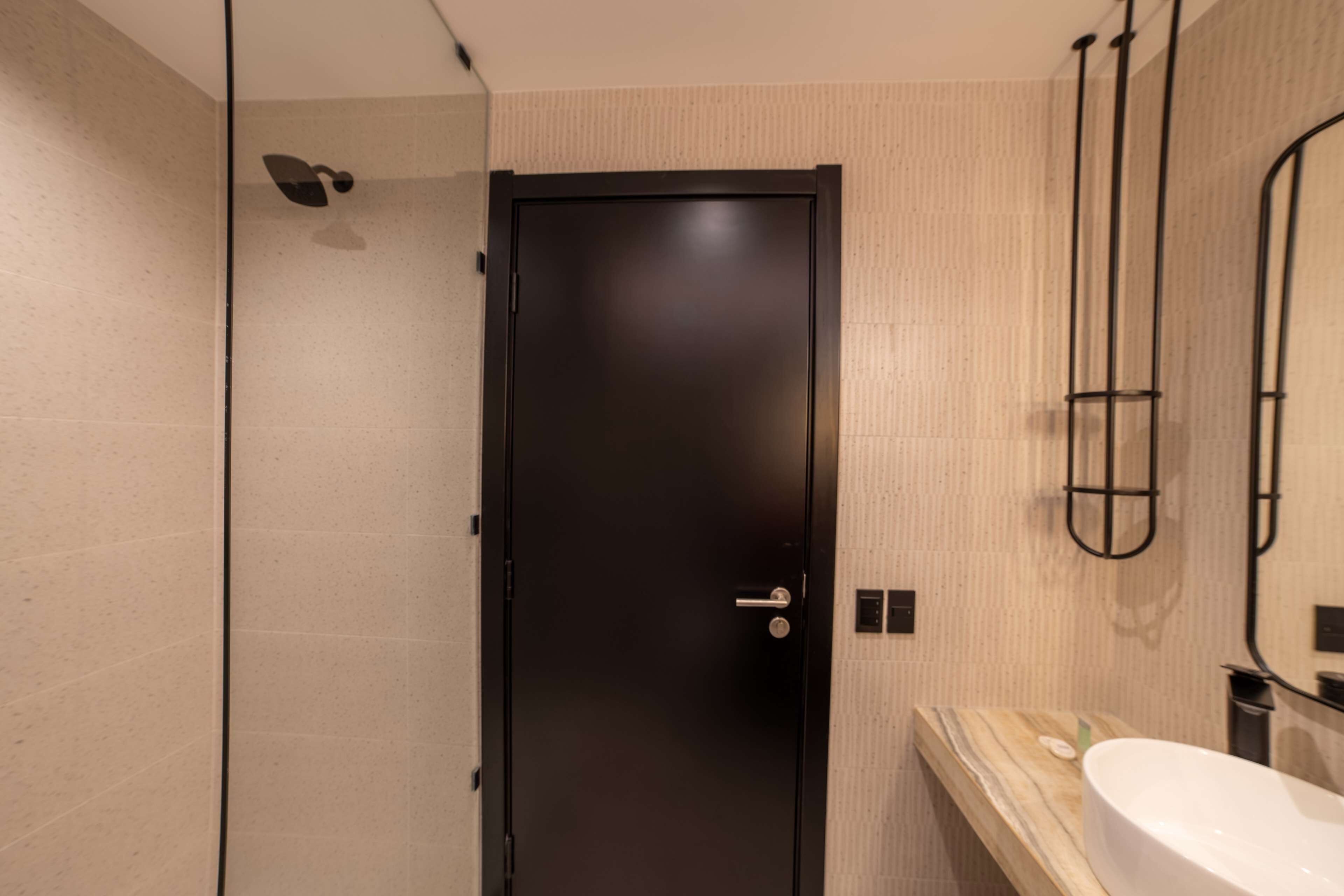 Un baño moderno con una puerta negra, un tocador de madera, un lavamanos blanco y un perchero para toallas.