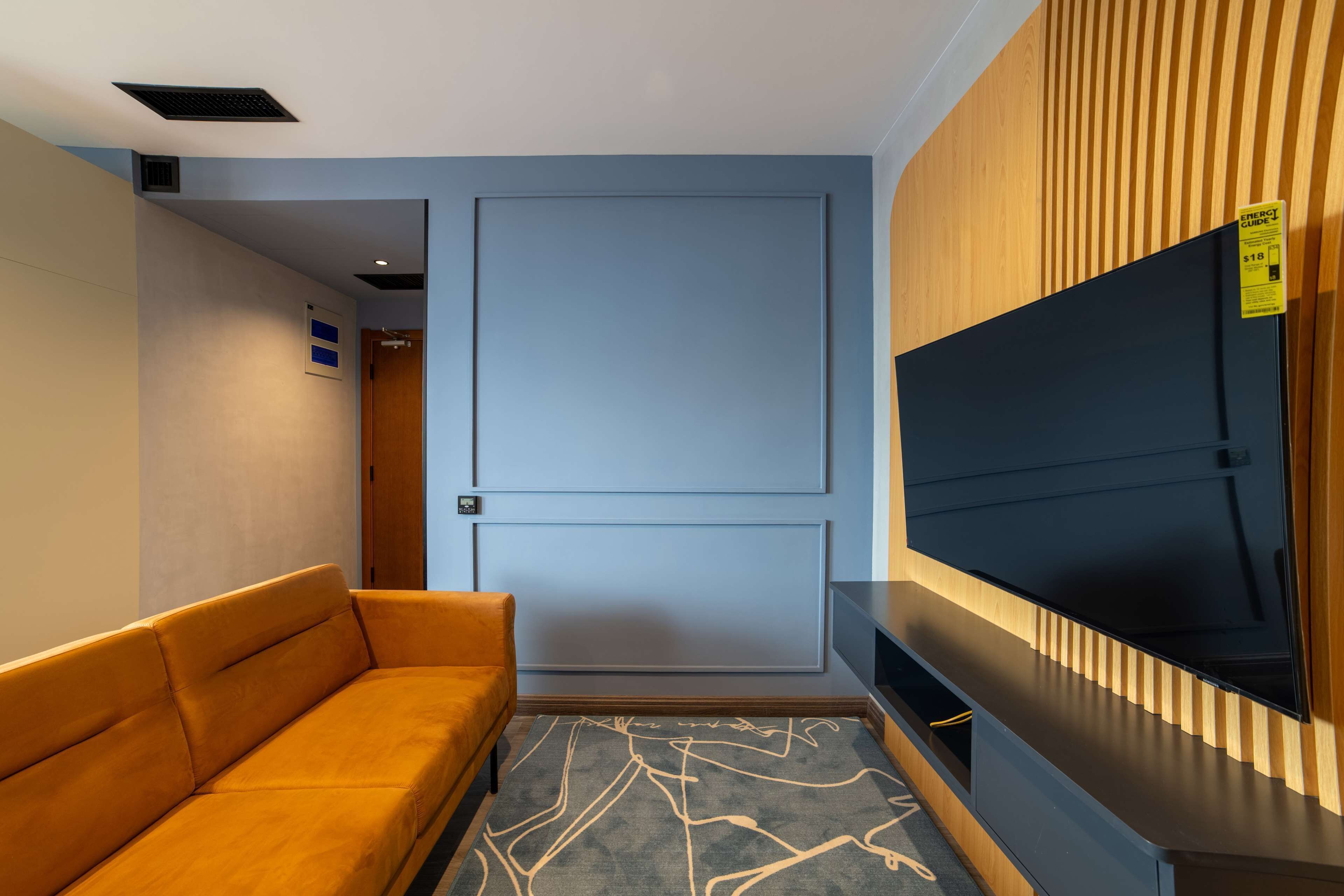 A modern hotel room with a sofa, a flat-screen TV mounted on a wooden panel, and a door with a light switch.