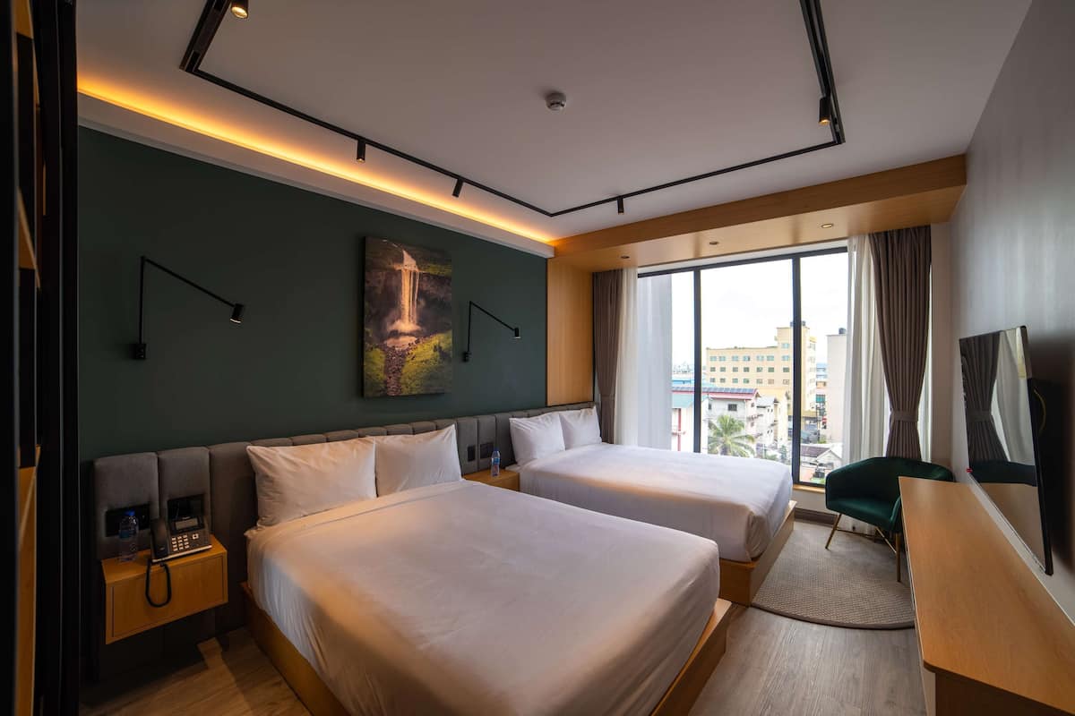 Ein Hotelzimmer mit zwei Betten, einem Schreibtisch, einem Stuhl, einem Gemälde, einem Telefon und einem Fenster mit Blick auf die Stadt.