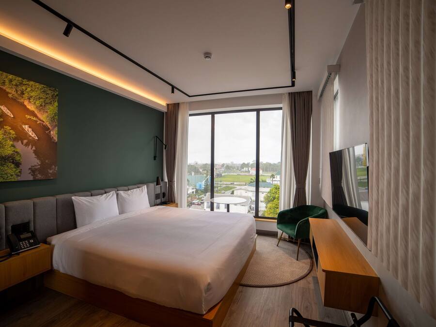 Ett modernt hotellrum med en stor säng, ett skrivbord, en stol, en TV och utsikt över staden genom stora fönster.