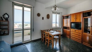 Een ruime woonkamer met een grote schuifpui naar een balkon, een eettafel met stoelen, houten kasten en een televisie.