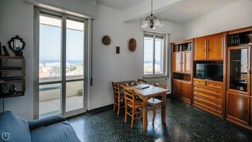 Un vaste espace de vie comprenant une grande porte-fenêtre coulissante donnant sur un balcon, une table à manger avec des chaises, des meubles en bois et un téléviseur.