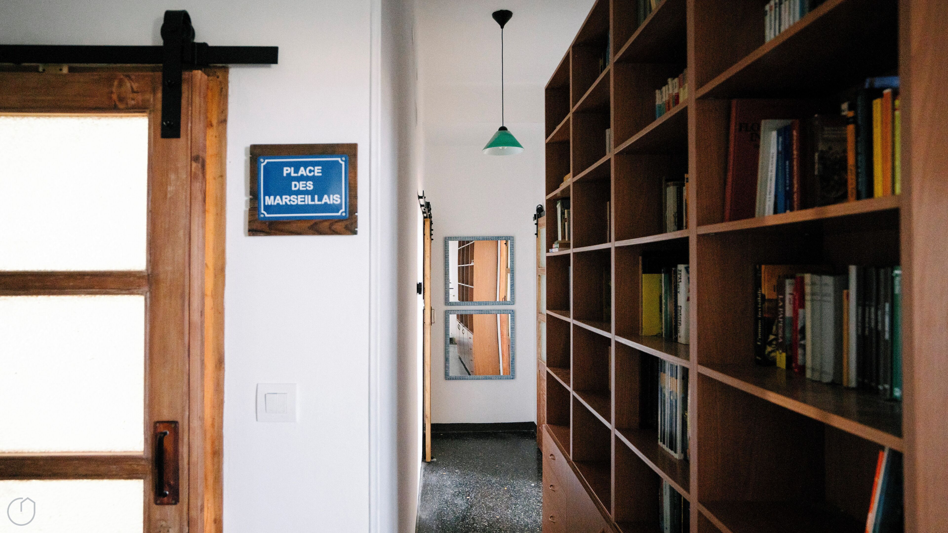 Un couloir avec une porte en bois, une enseigne indiquant « PLACE DES MARSEILLAIS », un luminaire suspendu vert et une bibliothèque remplie de livres.
