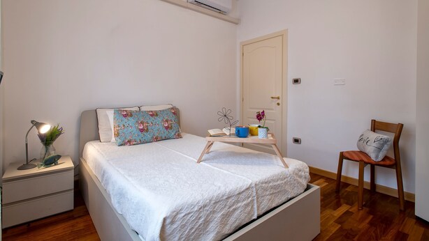 A bedroom with a bed, bedside table, chair, and an air conditioning unit. - Sardinia Re - 4 Mori Apartment (Sassari)