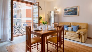 Un comedor con mesa de madera para cuatro personas, un sofá y un refrigerador en un apartamento moderno con balcón.