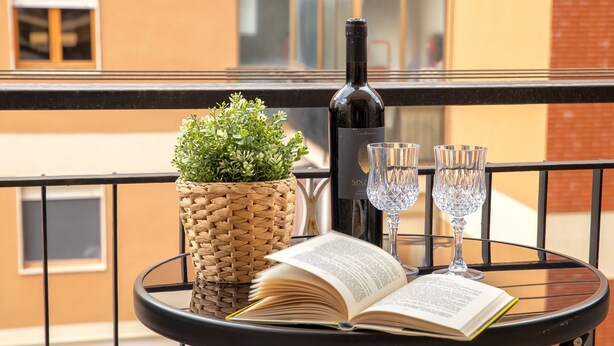 Una mesa redonda con un libro, una botella de vino, dos copas de vino y una cesta tejida con plantas verdes en un balcón.