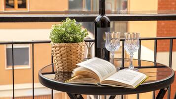 Une table ronde avec un livre, une bouteille de vin, deux verres à vin et un panier tressé contenant des plantes vertes, le tout sur un balcon.