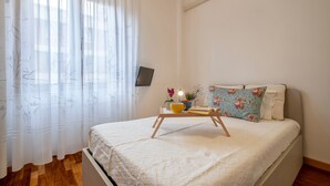 Una cama cuidadosamente hecha con un cojín floral, una mesita de noche con una taza y una planta pequeña, y cortinas de encaje con estampado floral.
