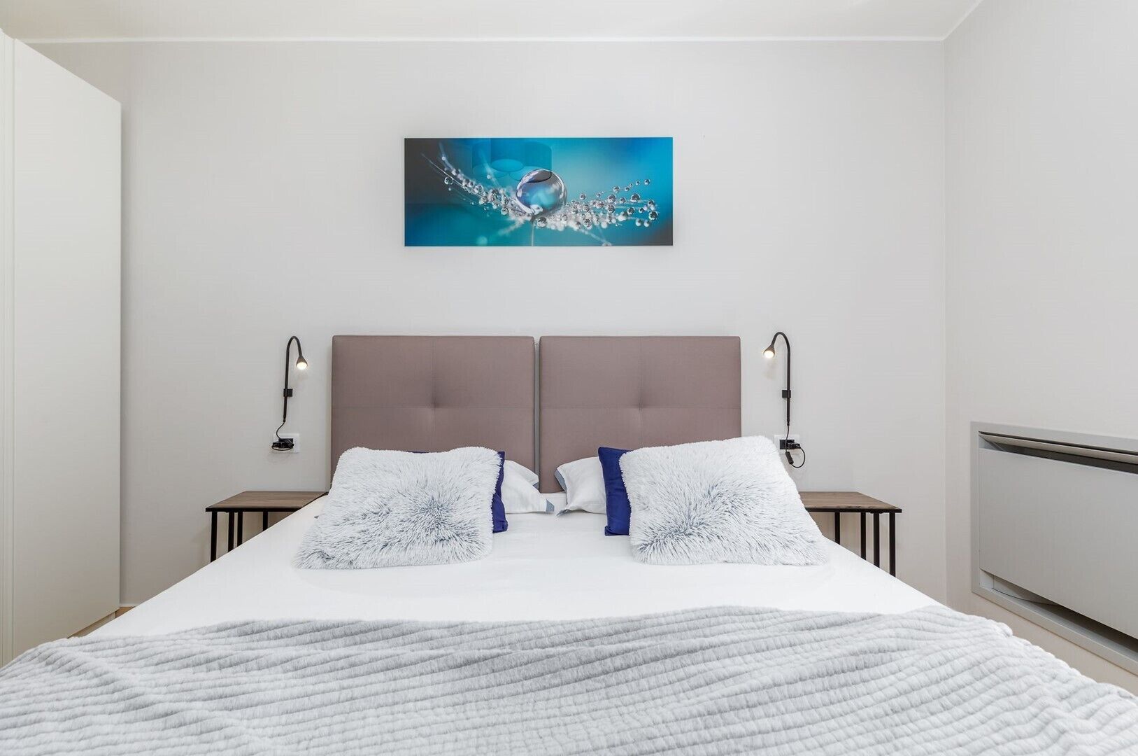 A modern bedroom with a large bed, two bedside tables, a headboard, and a wall-mounted artwork.