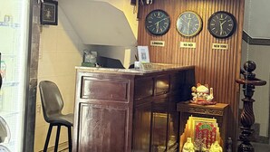 A hotel reception desk with clocks displaying London, Hanoi, and New York times, a small shrine with Buddha statues, and a wooden panel wall with the hotel's name and address. - Nhu Y Hotel (Ho Chi Minh City)