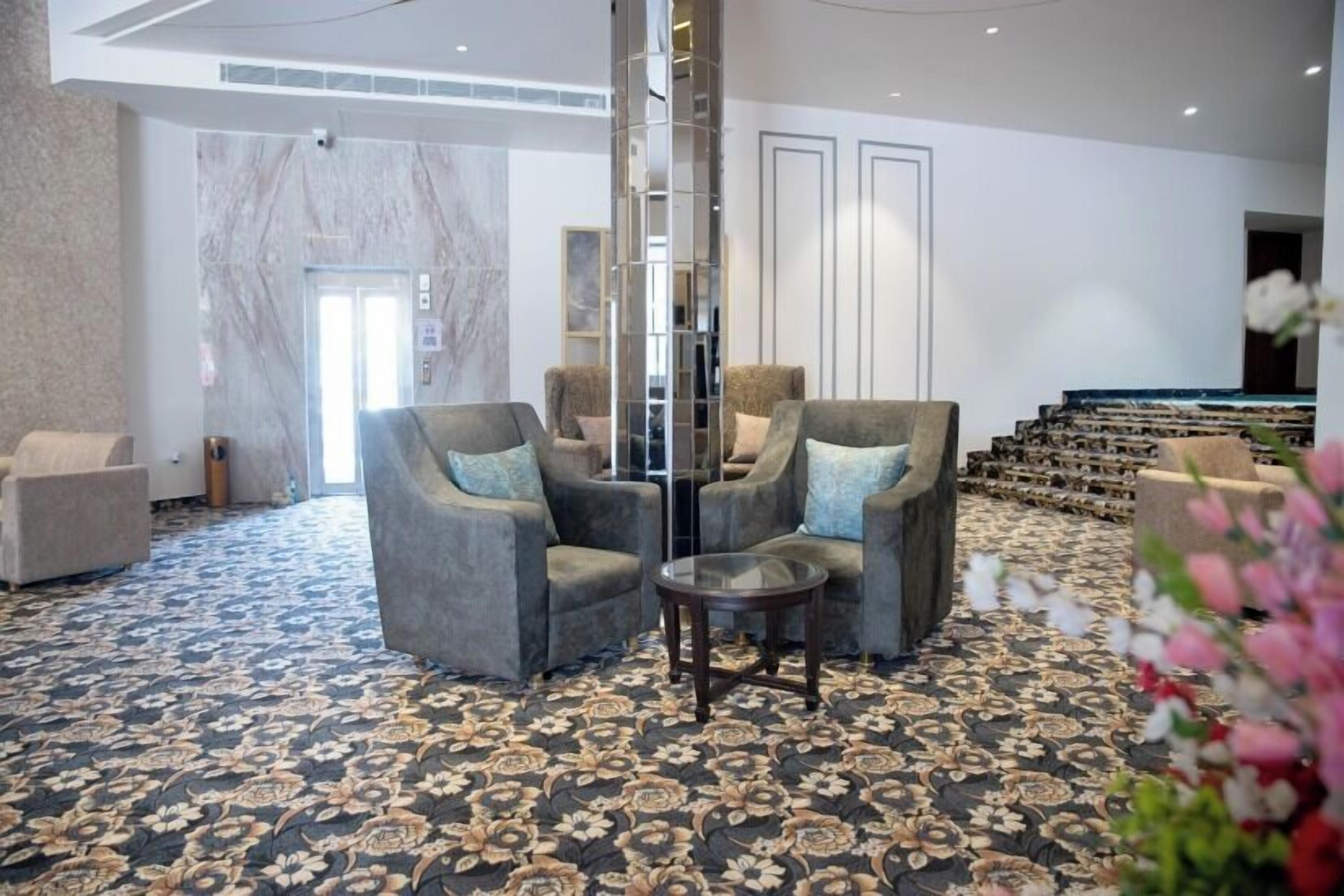 A hotel lobby with a patterned carpet, two grey armchairs with blue cushions, a small round table, and a tall, narrow mirror.