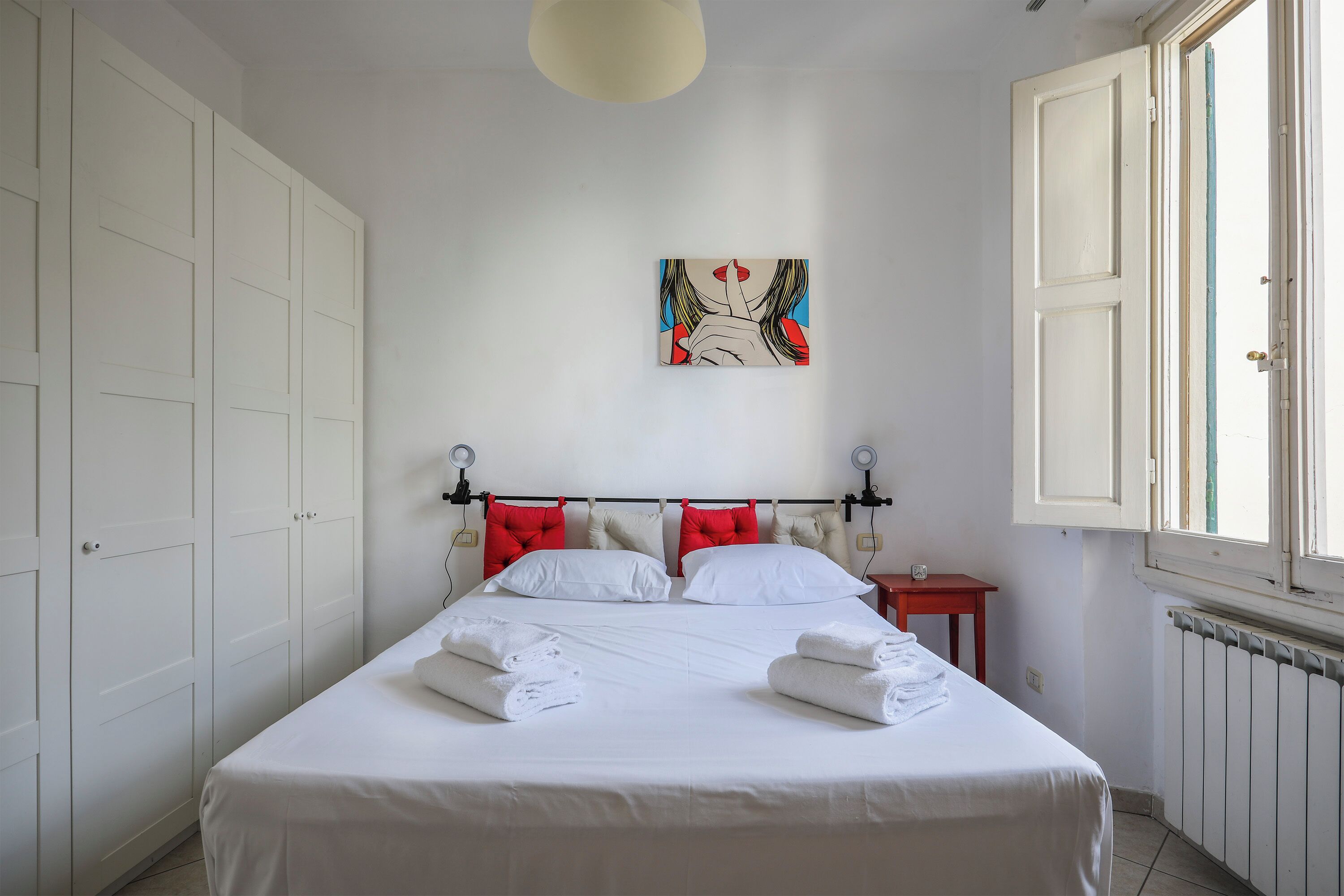 A neatly made bed with white linens and red pillows, a painting on the wall, and a window with open shutters.