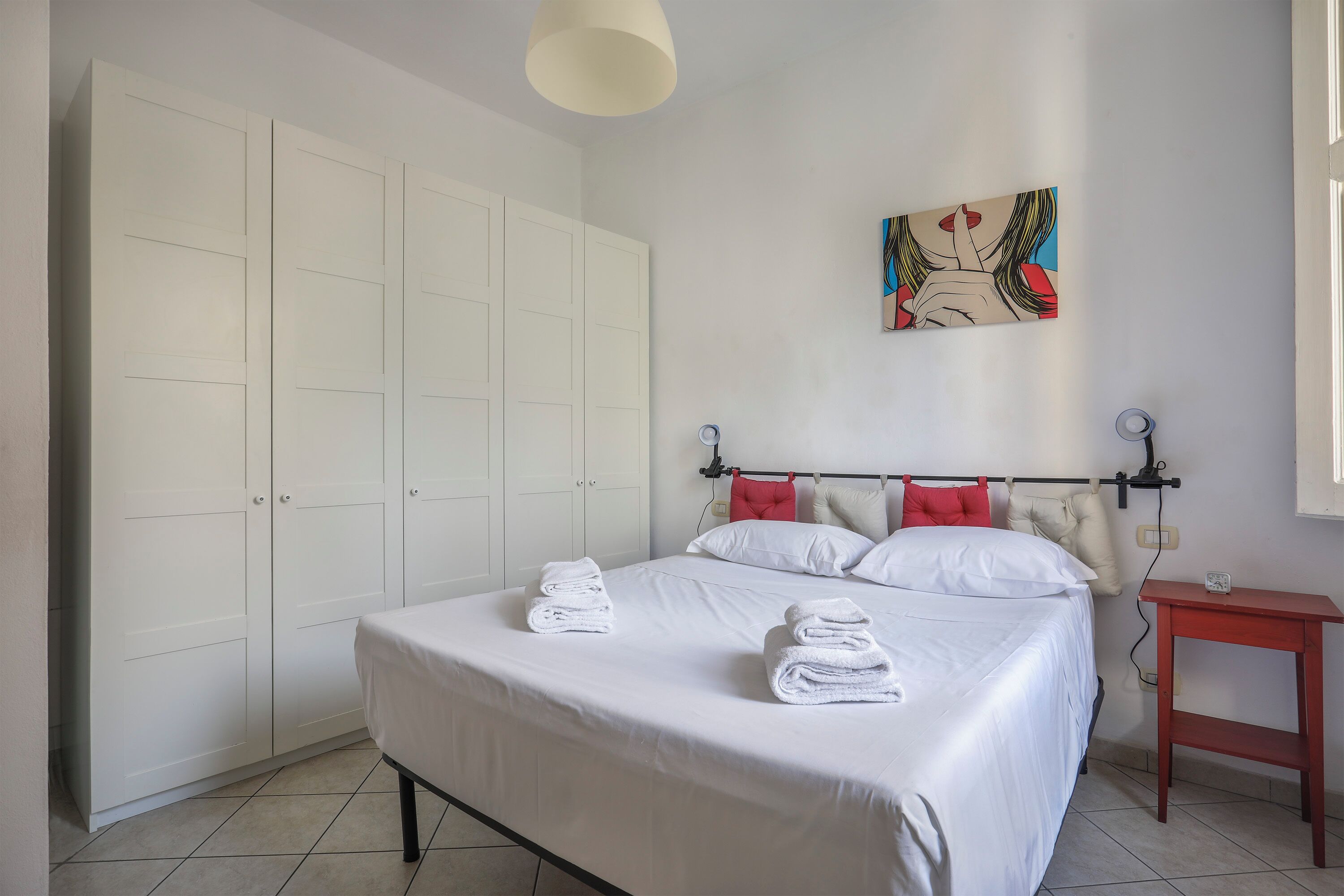 A bedroom with a bed, white curtains, a red bedside table, and a painting on the wall.
