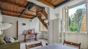 A room with a bed, a wooden ceiling, a window with a view of trees, and a small table with a lamp. - Angelico Urban Loft (Florence)
