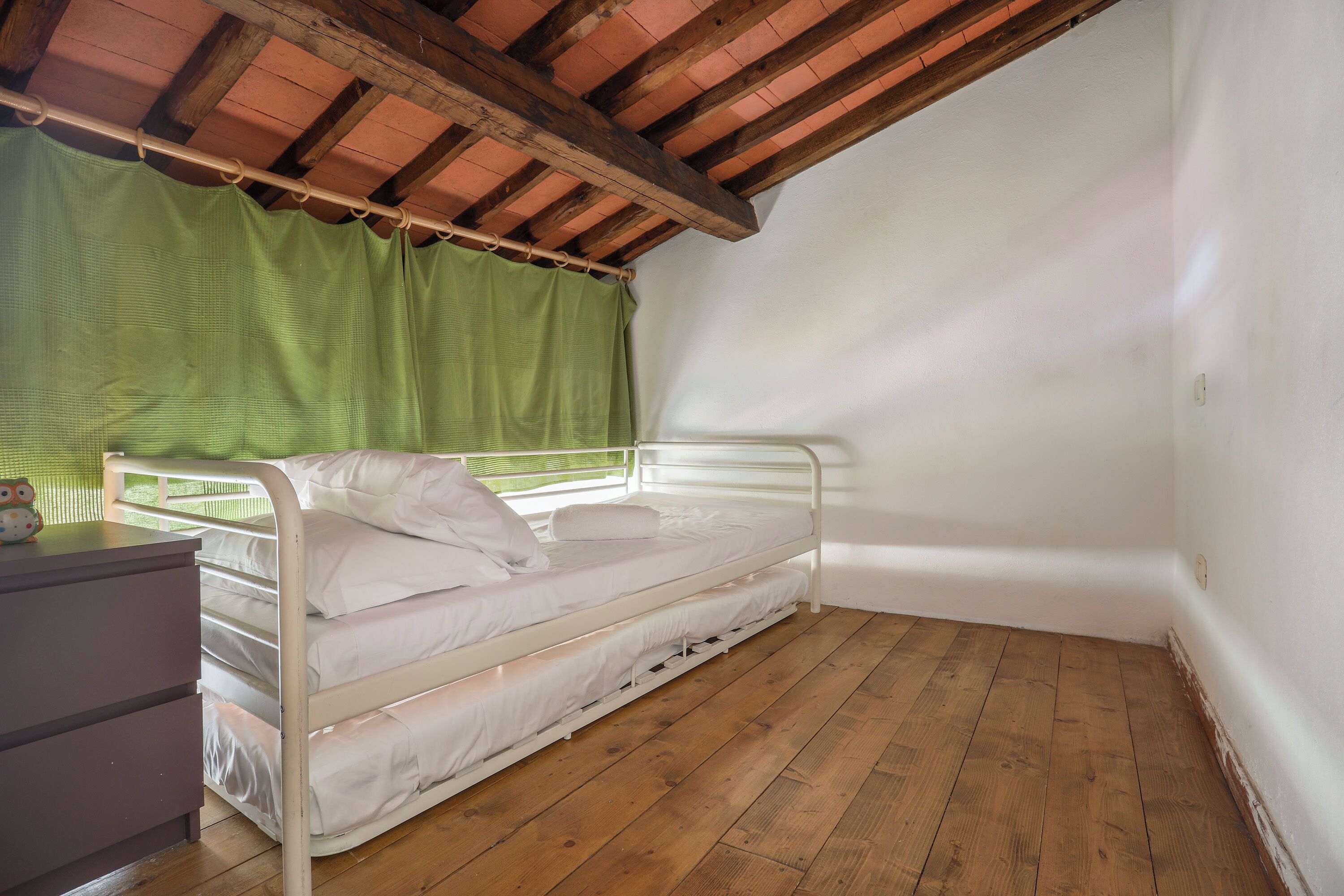 Une chambre avec un lit simple, un plancher en bois, un cadre de lit en métal, un rideau vert et un mur blanc.