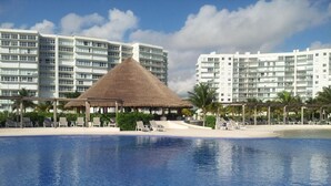 A swimming pool with a thatched-roof structure, surrounded by lounge chairs and palm trees, with modern buildings in the background.