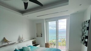 A bedroom with a bed, a ceiling fan, a large window with a view of the ocean, and a balcony.