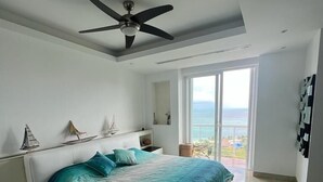 A bedroom with a bed, a ceiling fan, a nightstand, a lamp, and a view of the ocean through large windows.
