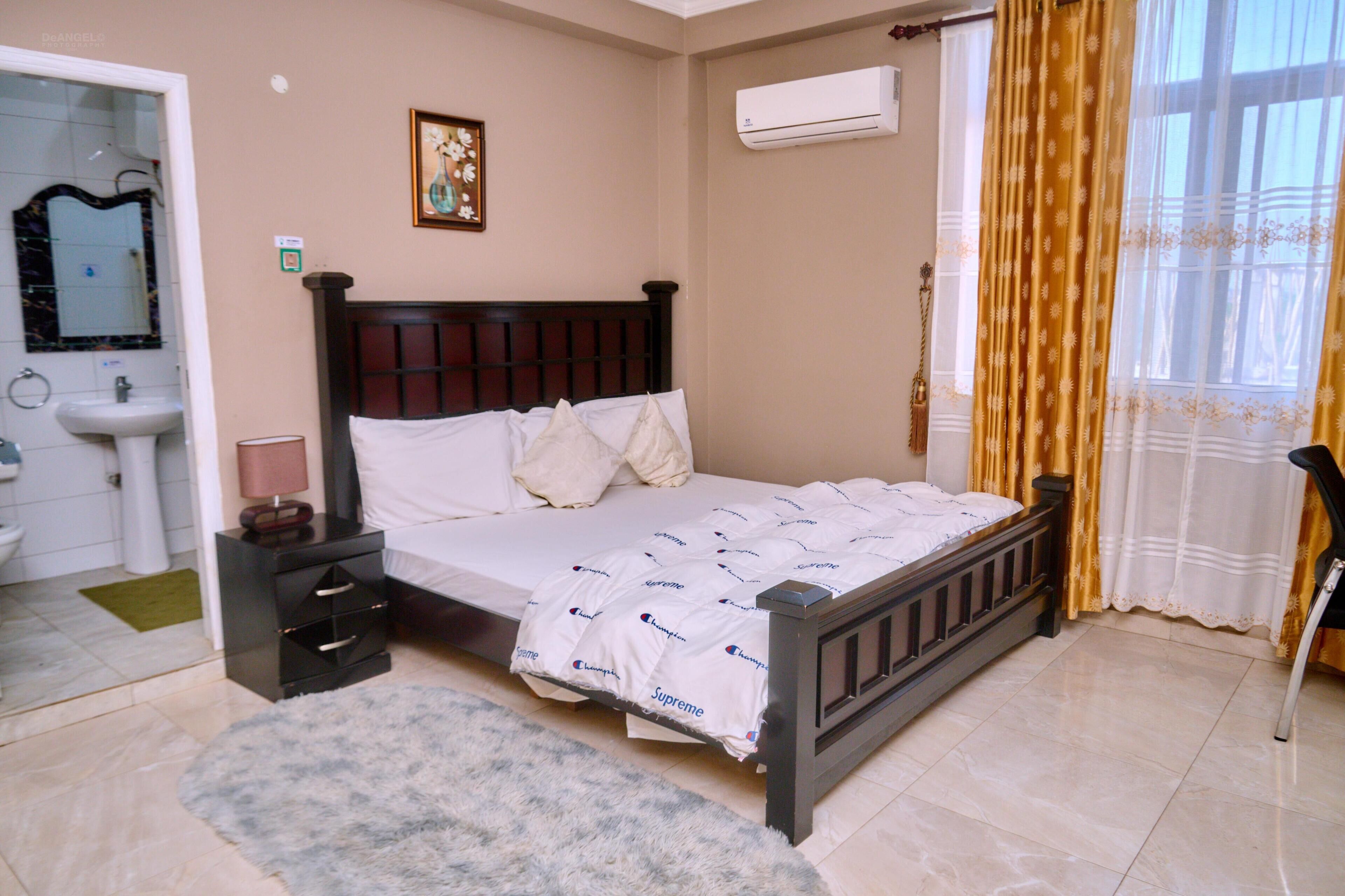 A bedroom with a bed, a nightstand, a bedside table, a mirror, a sink, and a window with curtains.