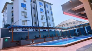A swimming pool area with a covered seating area, a building with multiple floors and balconies, and a clear sky. - Thy Favour Hotel (Sekondi-Takoradi)