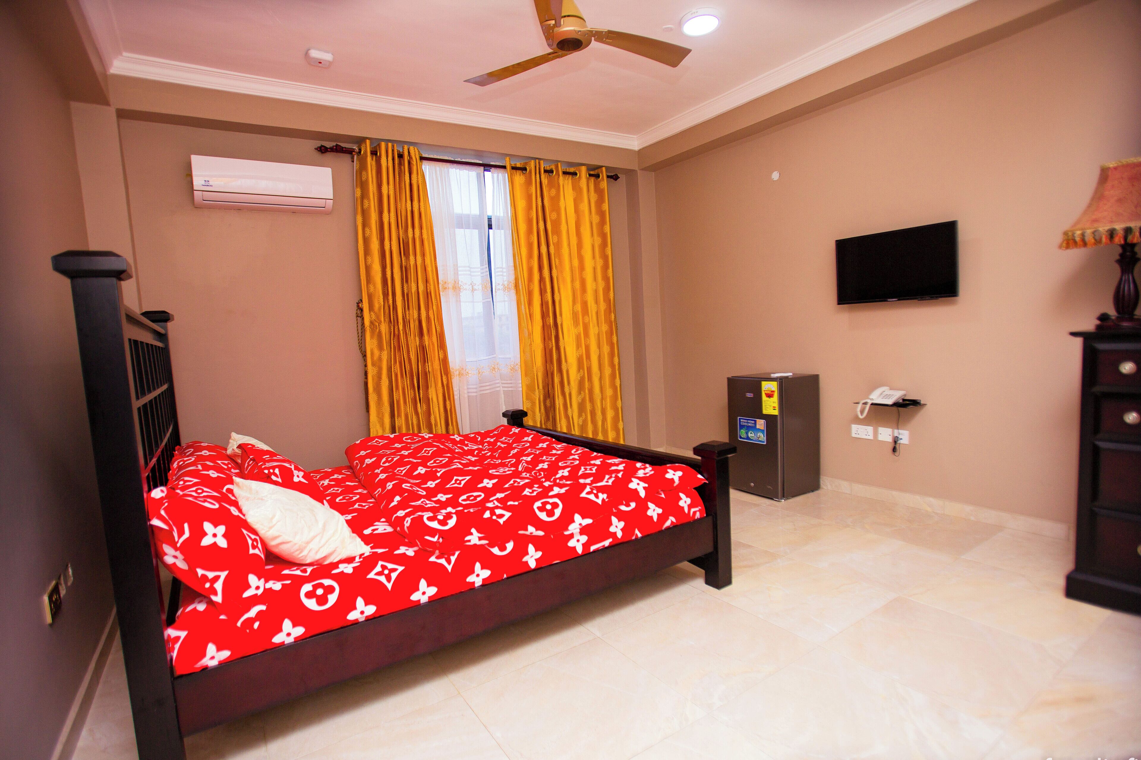 A bedroom with a bed, a ceiling fan, an air conditioning unit, a television, and a telephone.