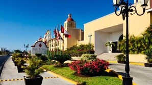 Entrada de un hotel con un letrero que dice "All Ritz-Carlton", varias banderas y un jardín bien cuidado con plantas y flores en macetas.