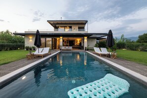 A modern two-story house with a swimming pool, outdoor seating, and a view of the mountains.
