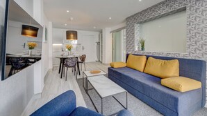 Una sala de estar moderna con un sofá azul y cojines amarillos, un área de comedor con una mesa y sillas, y una cocina al fondo.