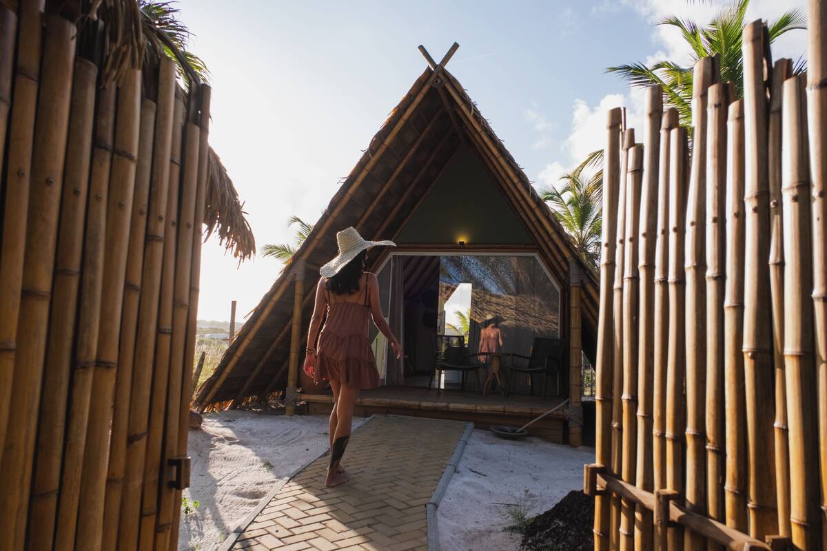 Une structure à ossature en bambou, surmontée d’un toit triangulaire, avec une entrée pavée et une personne portant un chapeau.